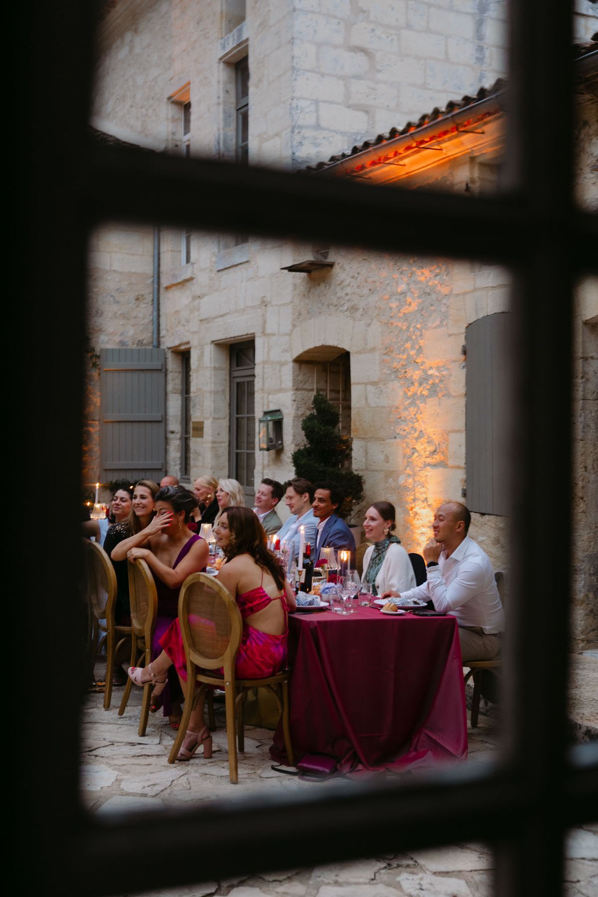 Outdoor reception in the stone patio courtyard at Domaine de Perrotin, with long tables dressed in raspberry-red linen, gold cane chairs and colourful florals for Rooa and Mark's wedding