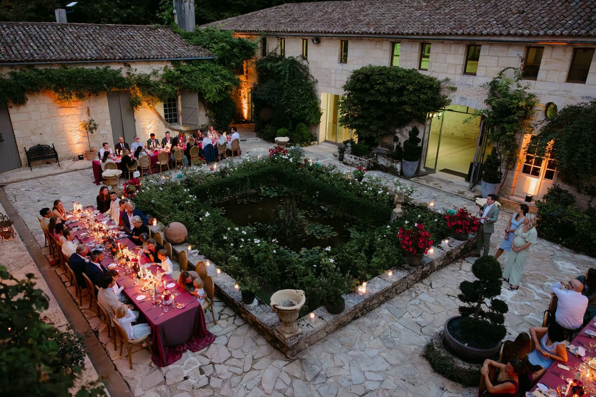 Outdoor reception in the stone patio courtyard at Domaine de Perrotin, with long tables dressed in raspberry-red linen, gold cane chairs and colourful florals for Rooa and Mark's wedding