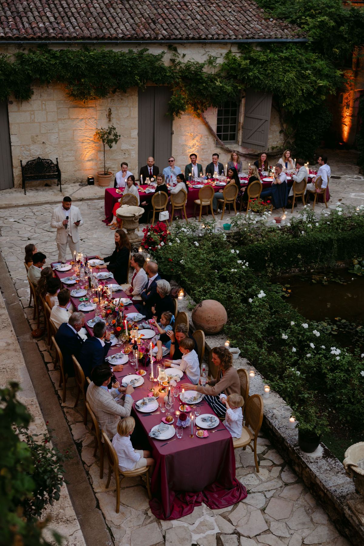 Outdoor reception in the stone patio courtyard at Domaine de Perrotin, with long tables dressed in raspberry-red linen, gold cane chairs and colourful florals for Rooa and Mark's wedding