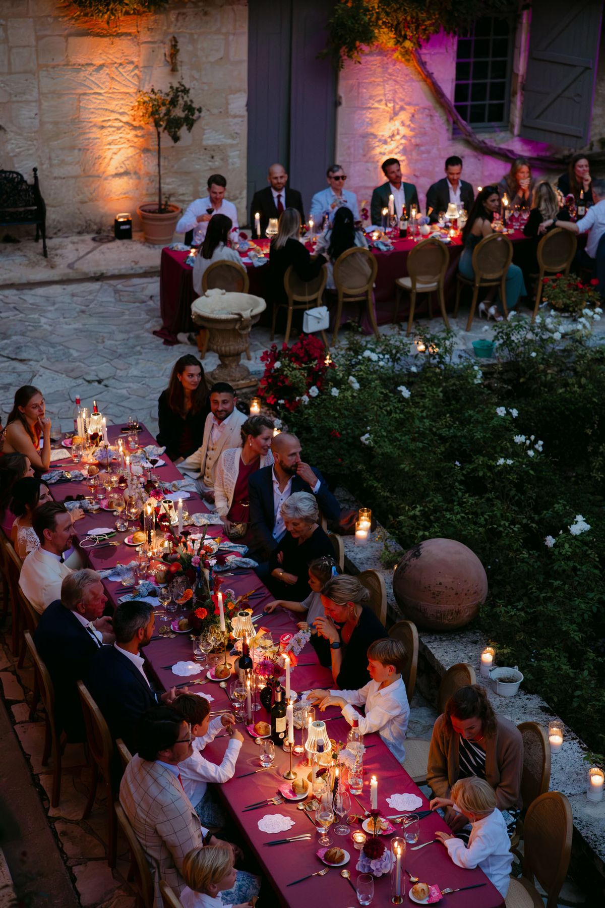 Outdoor reception in the stone patio courtyard at Domaine de Perrotin, with long tables dressed in raspberry-red linen, gold cane chairs and colourful florals for Rooa and Mark's wedding