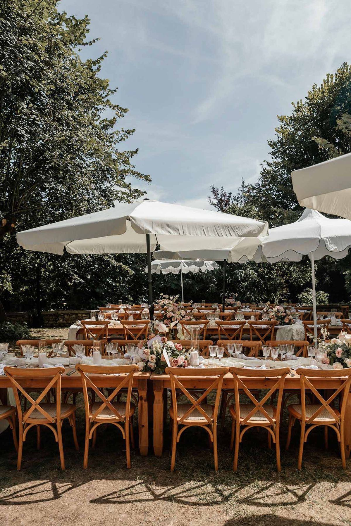 Outdoor reception setup with long wooden farm tables, cross-back chairs, and blush rose centerpieces under white umbrellas