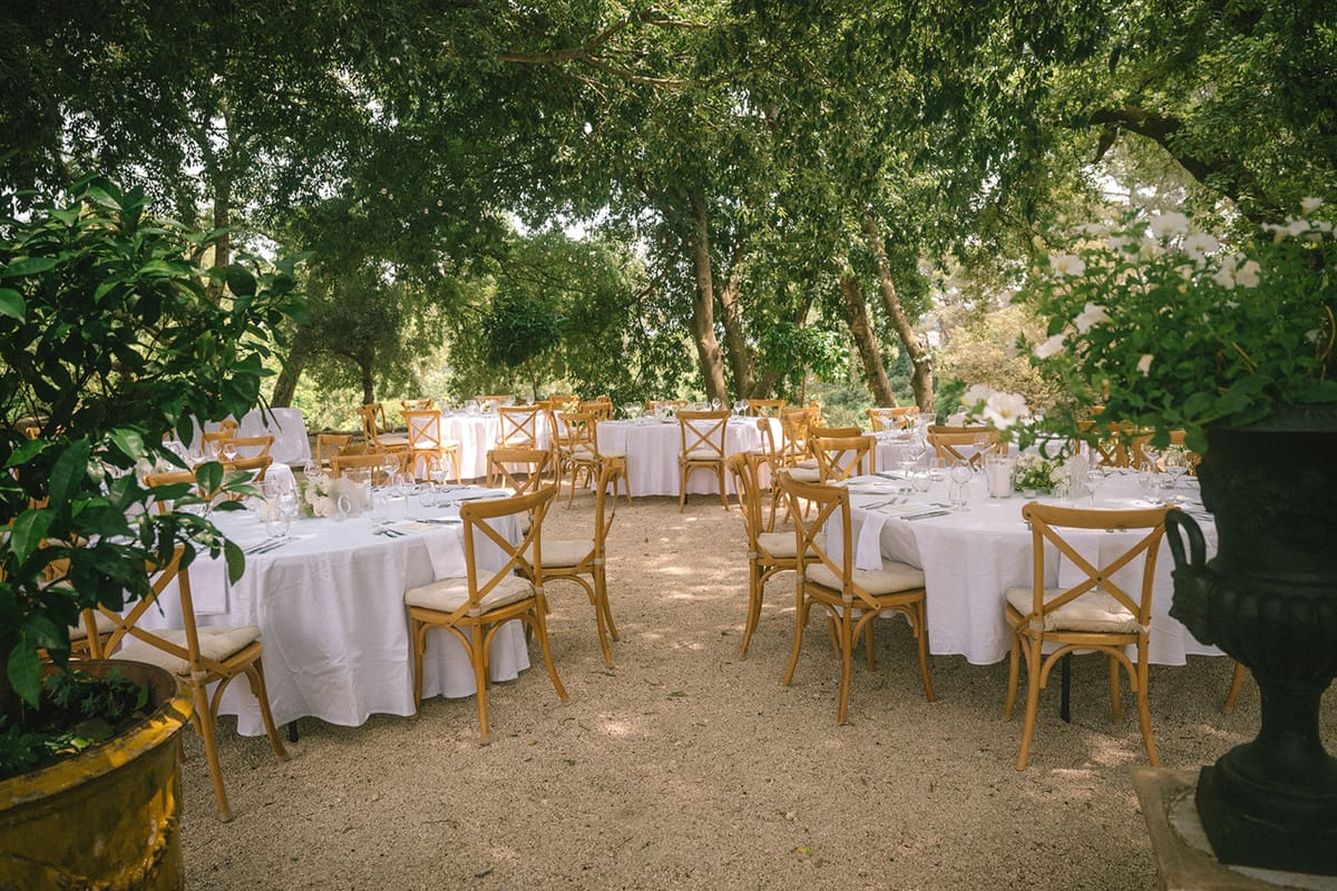 Outdoor reception setup with round white-clothed tables and cross-back chairs beneath mature trees on gravel garden
