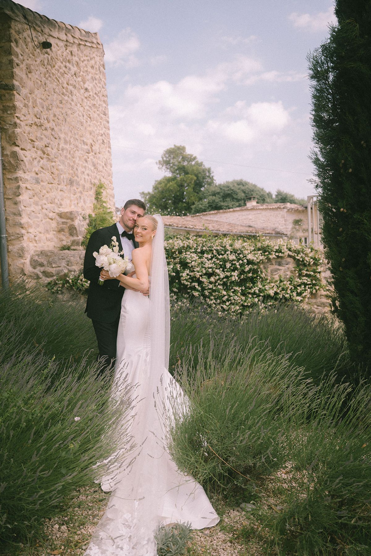 Bride and groom embracing among lavender rows at a stone French property with cypress trees and climbing roses