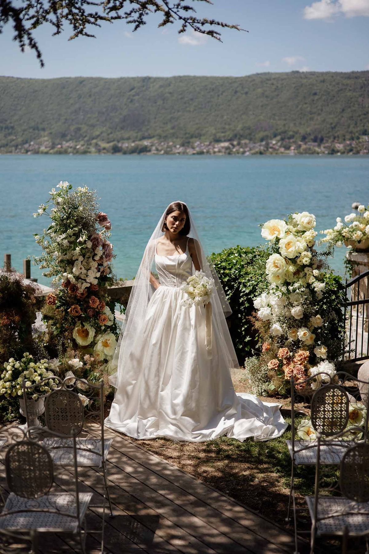 Bride in satin ball gown and veil standing between floral installations at lakeside ceremony with mountain view