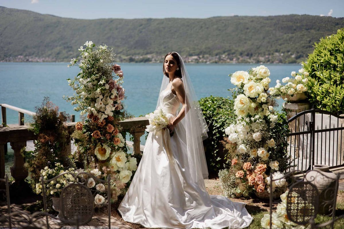 Bride in white satin ballgown with cathedral veil on lakeside terrace with terracotta and ivory rose stone urn arrangements