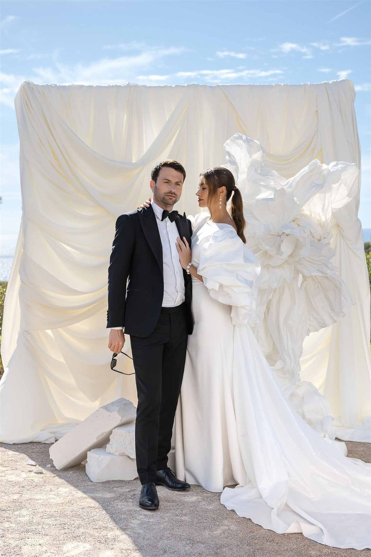 Couple before draped ivory backdrop with sculptural white florals, bride in puff-sleeve gown, coastal view behind
