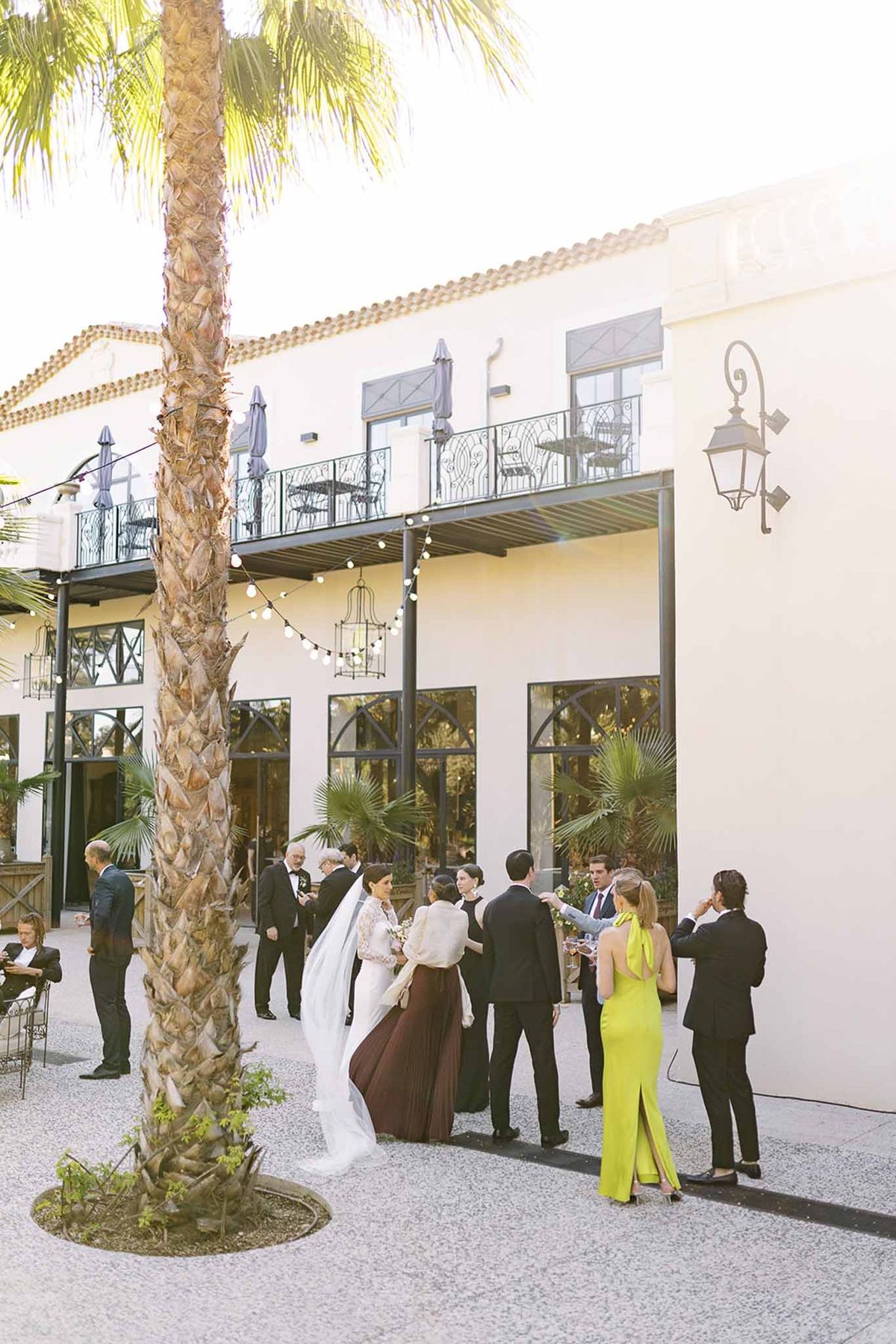 Golden hour cocktail gathering beneath wrought-iron balcony of white venue with string lights
