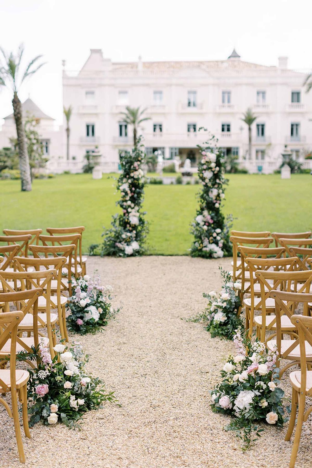 Pre-ceremony aisle with cross-back chairs, blush and white florals, and tall green columns at manor