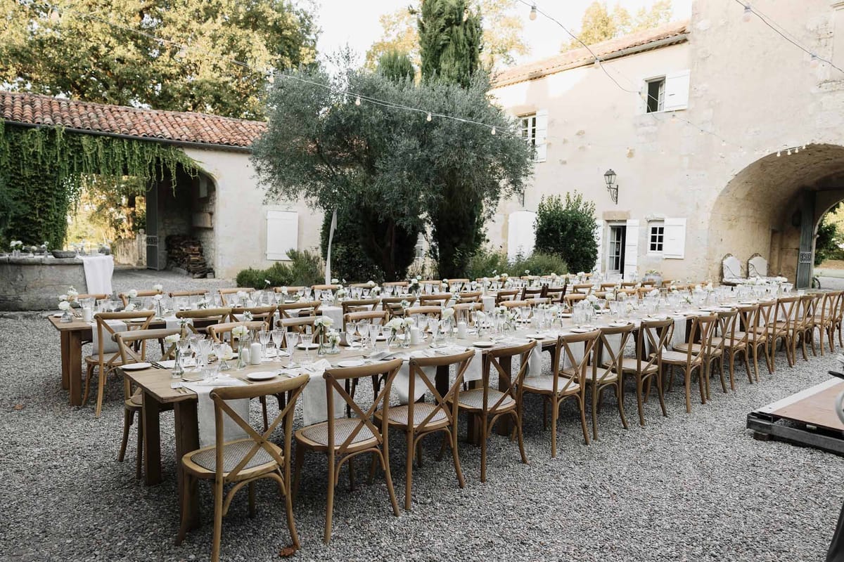 Outdoor reception setup in a Provencal courtyard with long farm tables, cross-back chairs, white florals, and overhead fai...