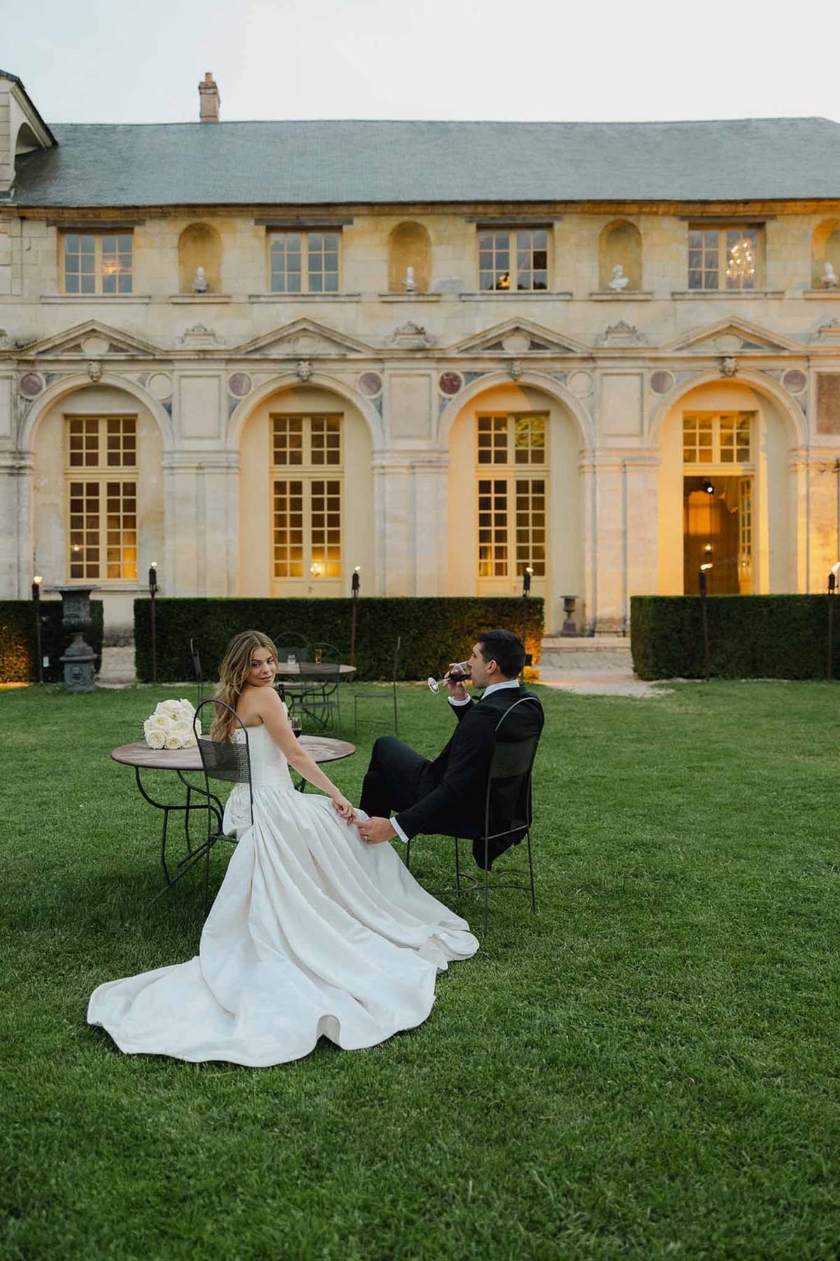 Bride and groom at bistro table on chateau lawn at dusk with warm interior light behind