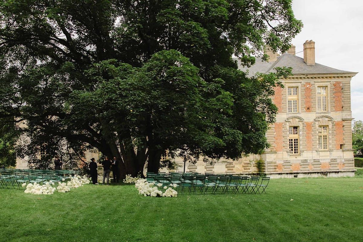Teal folding chairs on lawn with white hydrangea aisle clusters before brick-and-stone chateau
