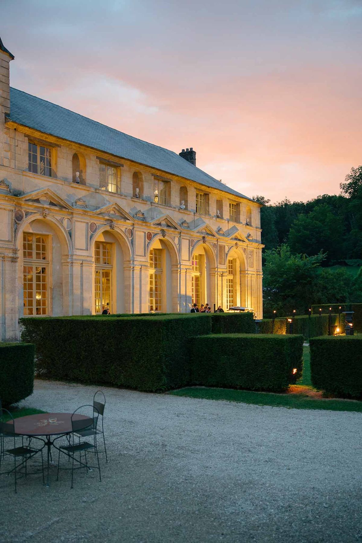 Classical stone chateau at dusk with warm uplighting, sculpted busts, boxwood hedges, and candlelit gravel pathways