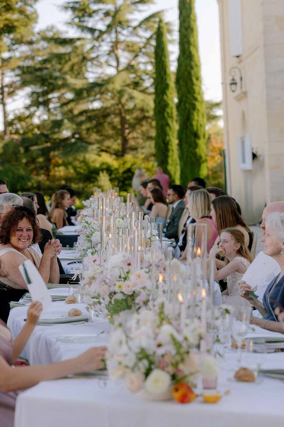 Long candlelit table with blush and cream rose runner beside chateau facade with 20 seated guests