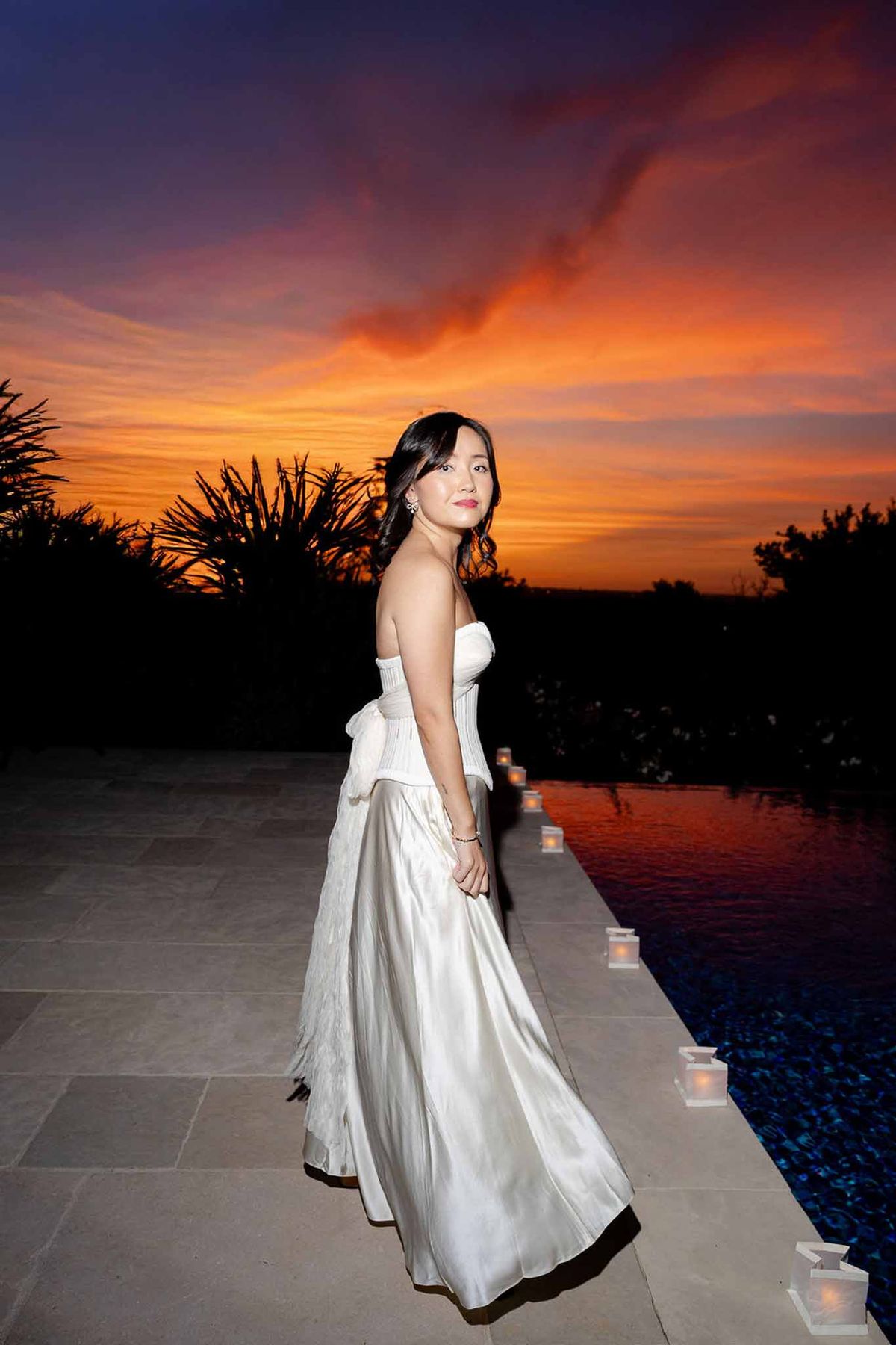 Bride in strapless satin gown poses beside illuminated pool at dusk with orange and purple sunset sky