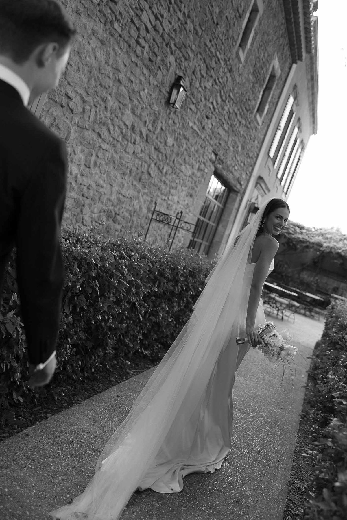 Black-and-white portrait of bride in strapless gown with cathedral veil looking back at groom along chateau pathway
