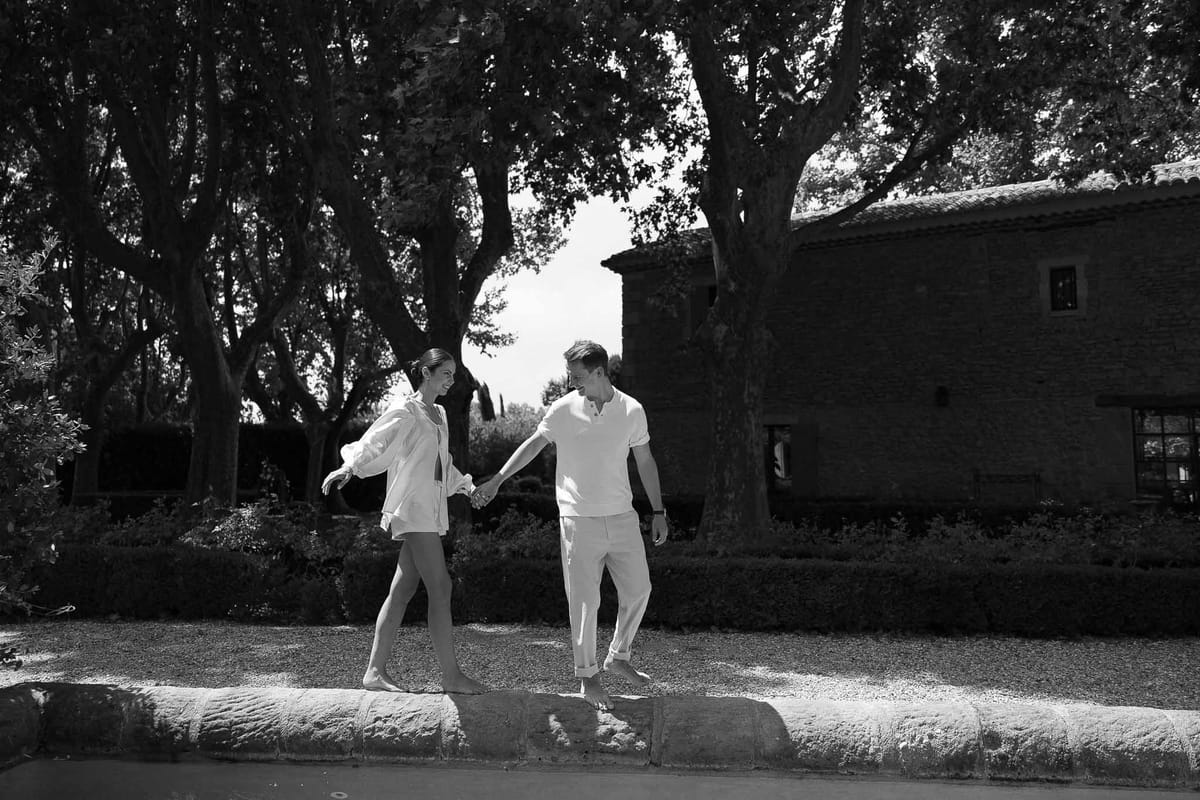 Couple in all-white casual outfits laughs balancing barefoot on stone pool edge at French estate in B&W