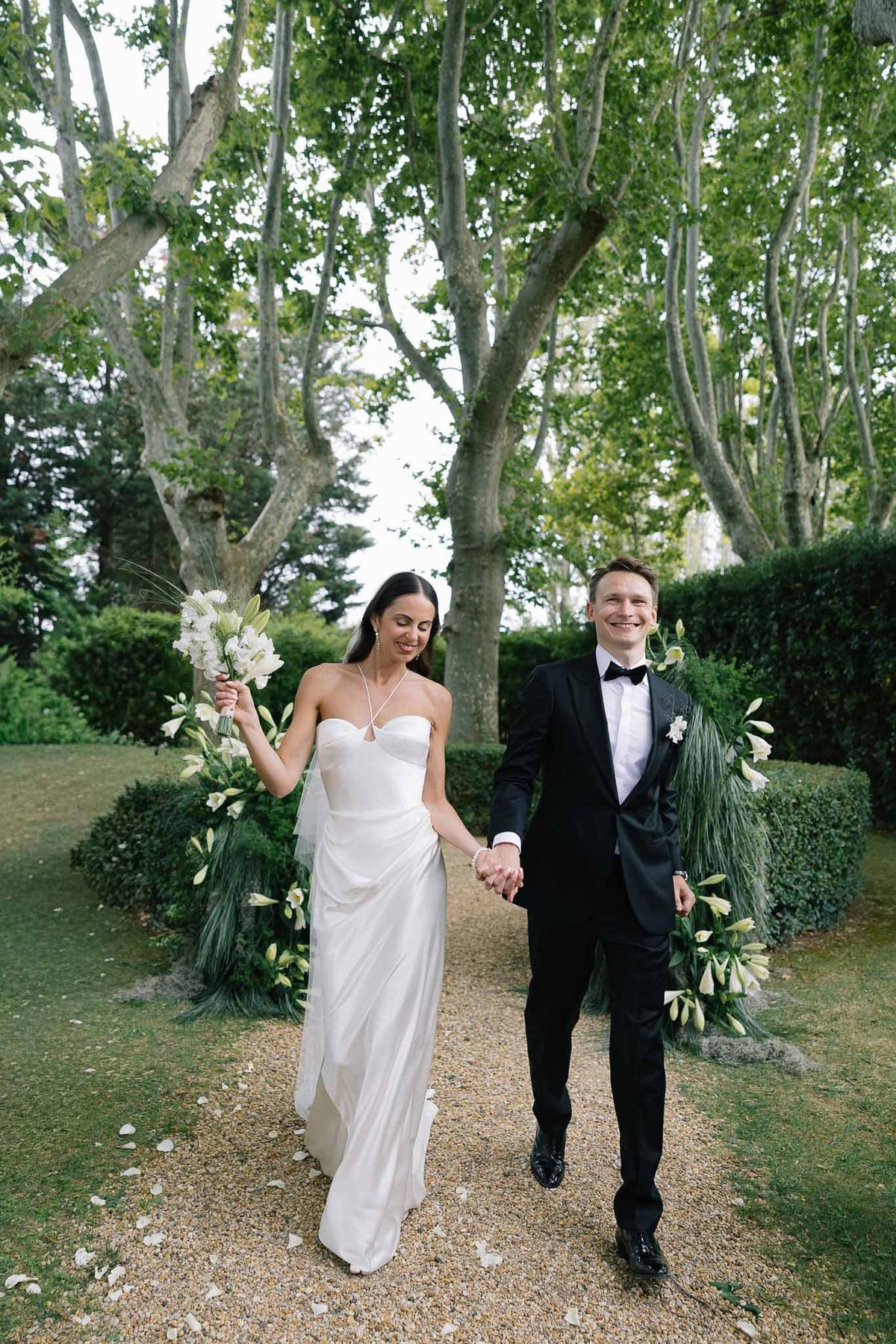 Bride and groom walk hand-in-hand along petal-strewn path flanked by tall white lily floral installations