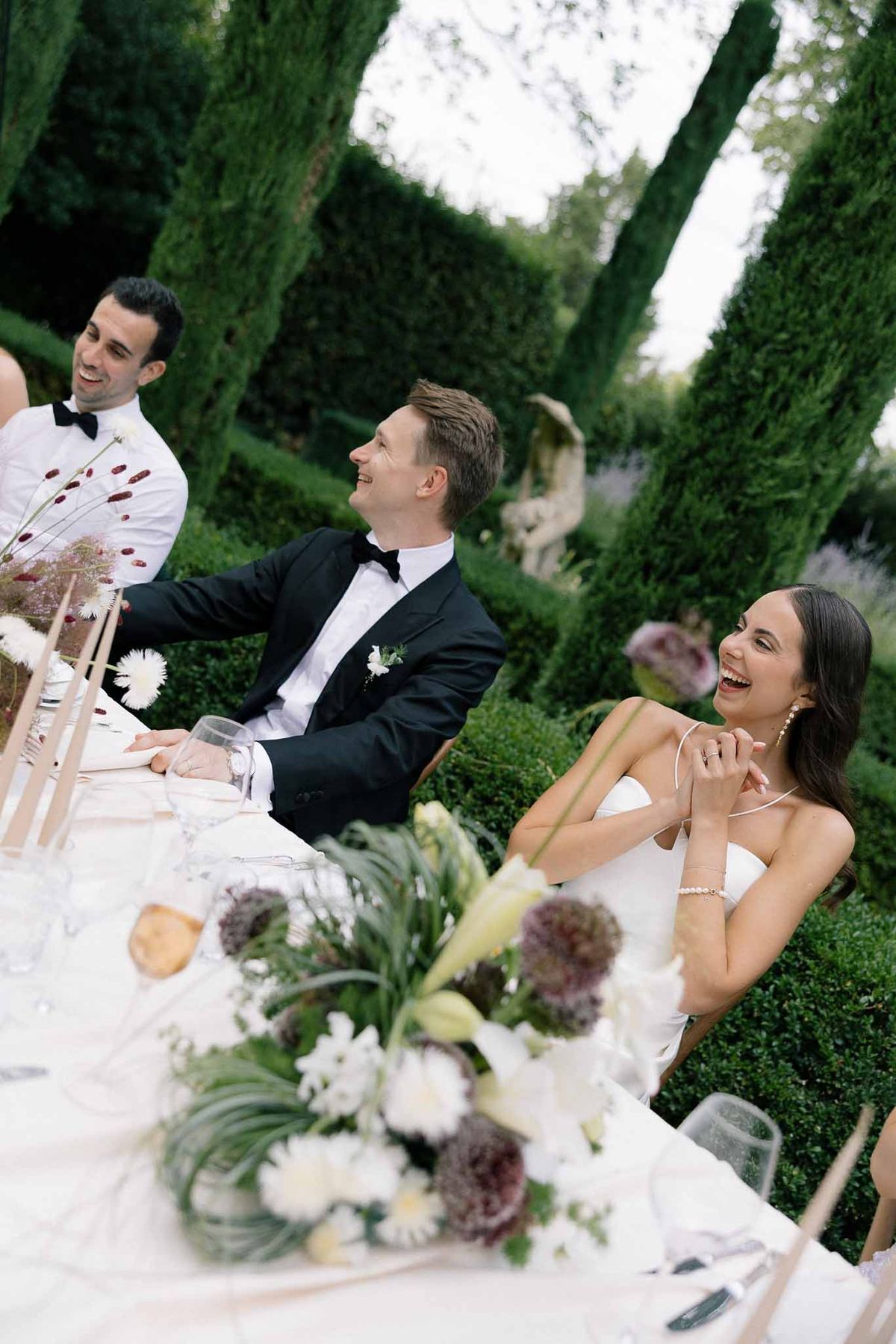 Bride and groom laughing at head table with burgundy allium and white gerbera centerpieces in formal garden
