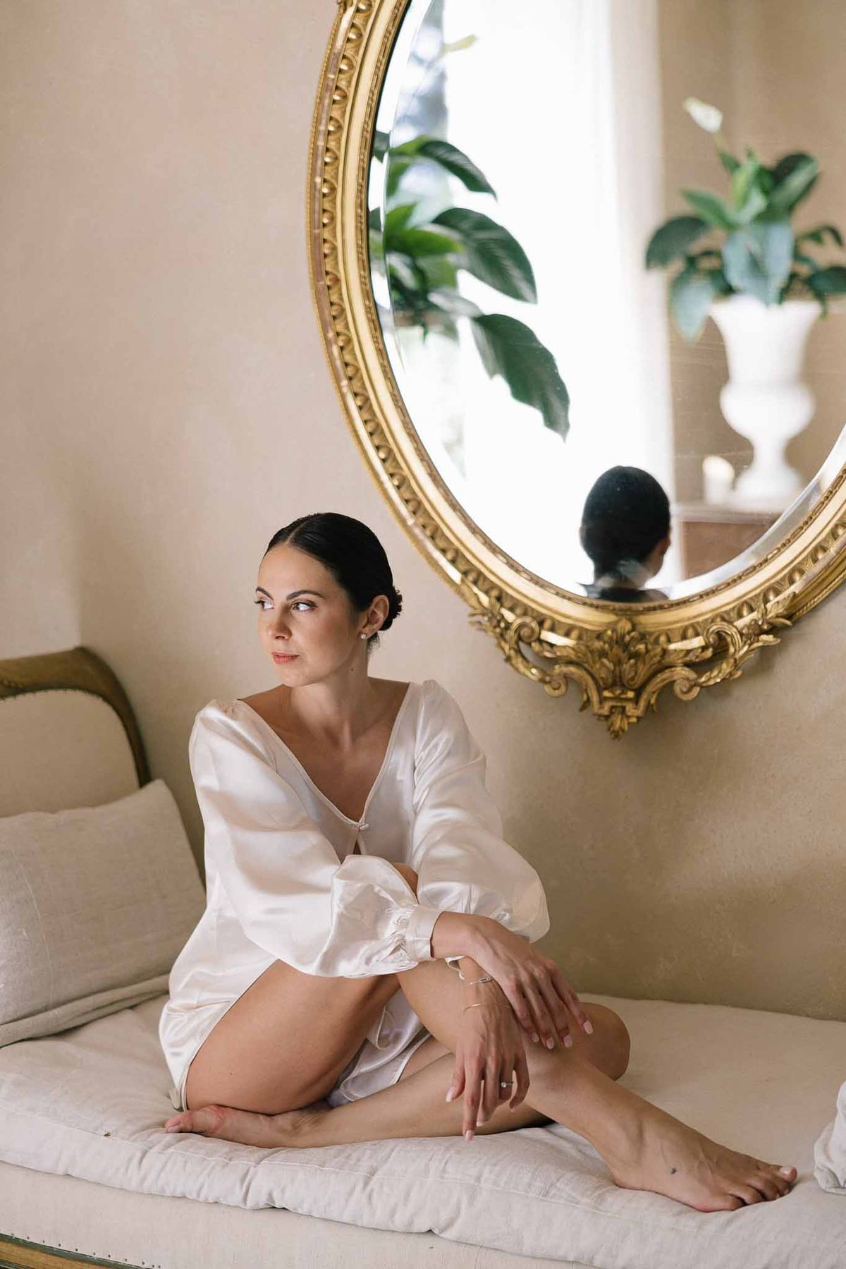 Bride in white puff-sleeve satin robe seated on daybed with gold oval mirror and tropical leaves