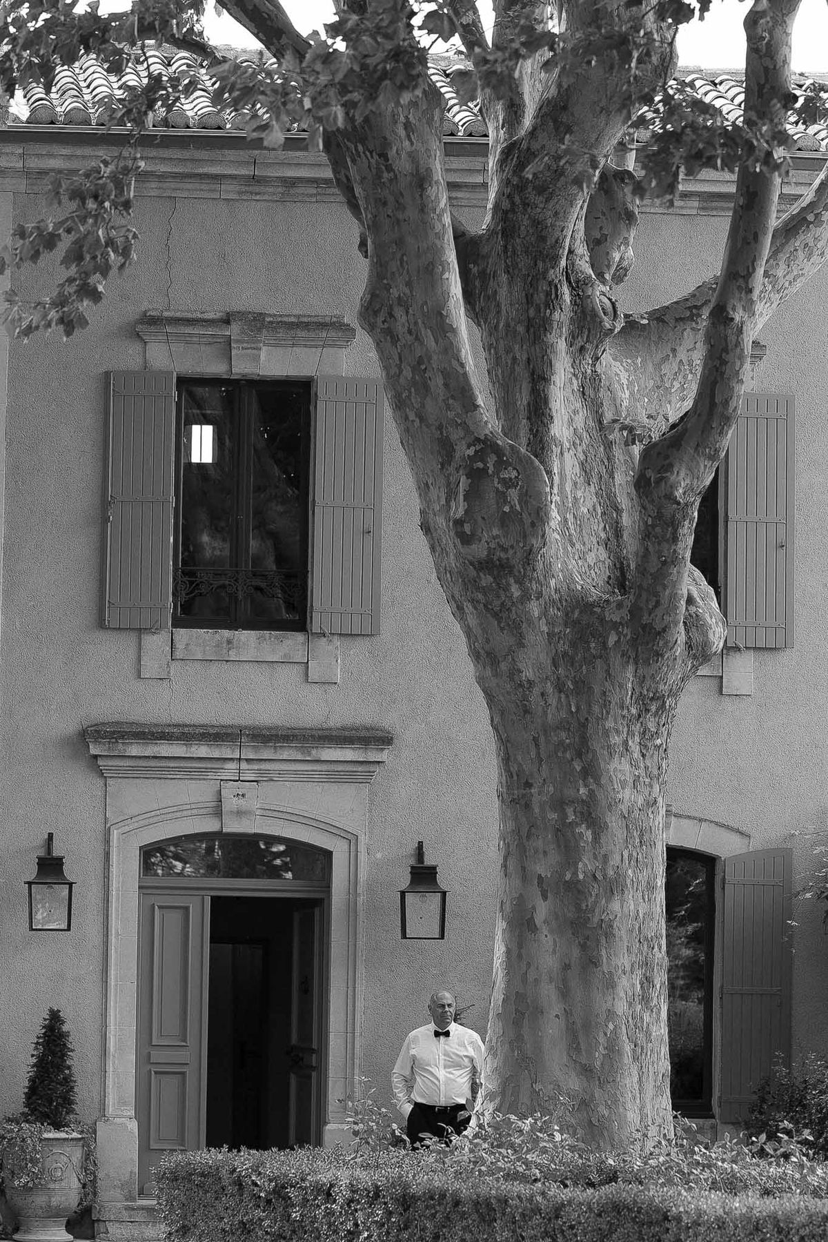 Black and white wide shot of groom standing beneath plane tree in front of Provencal mas with arched doorways