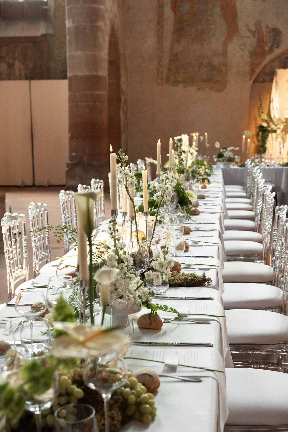 Long banquet table with calla lily and fern runner, silver candelabras, and ghost chairs in frescoed venue