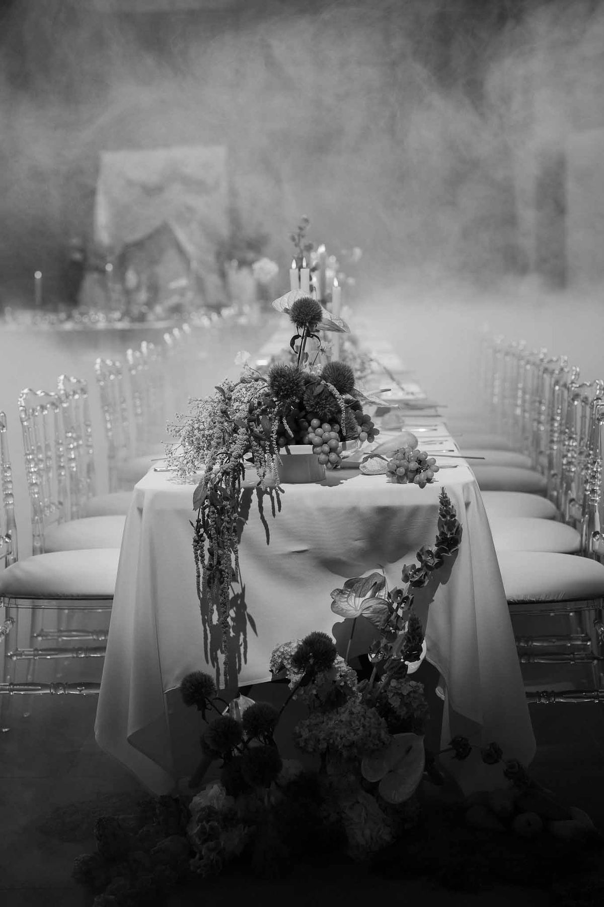 Black-and-white view down a long banquet table with trailing amaranthus, thistles, grapes, and taper candles