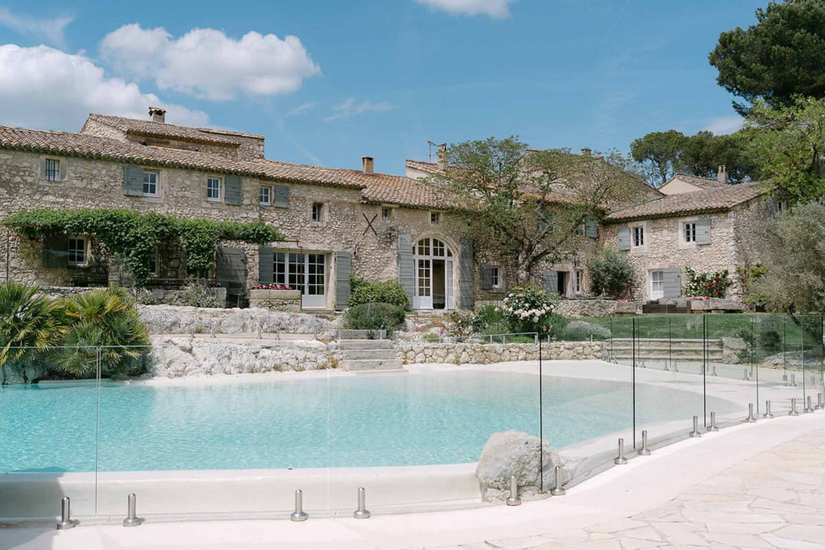 Provencal stone mas with grey-blue shutters, glass pool fence, terraced retaining walls, and pink roses