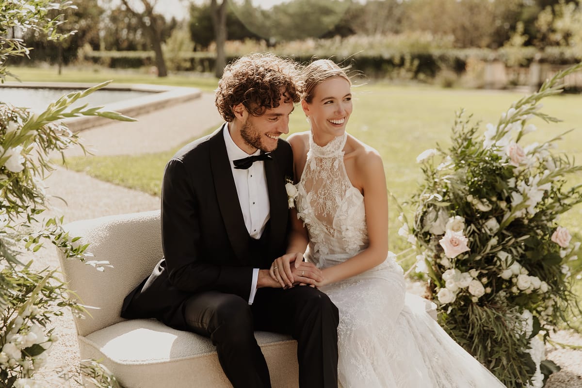 Bride and groom laughing on cream sofa framed by blush and white floral arrangements in garden