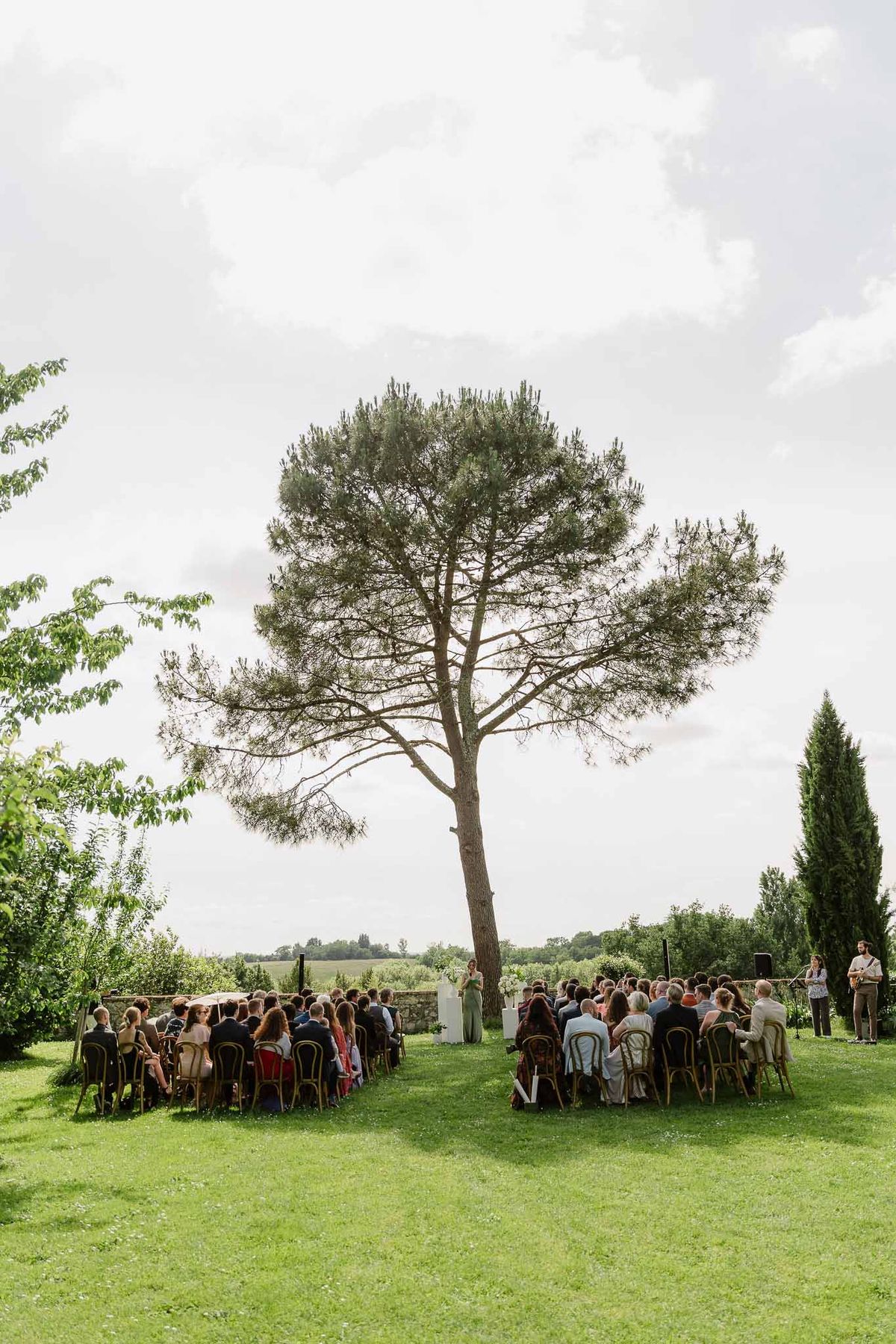Ceremony under umbrella pine on lawn with bistro chairs, acoustic guitarist, and countryside beyond stone wall