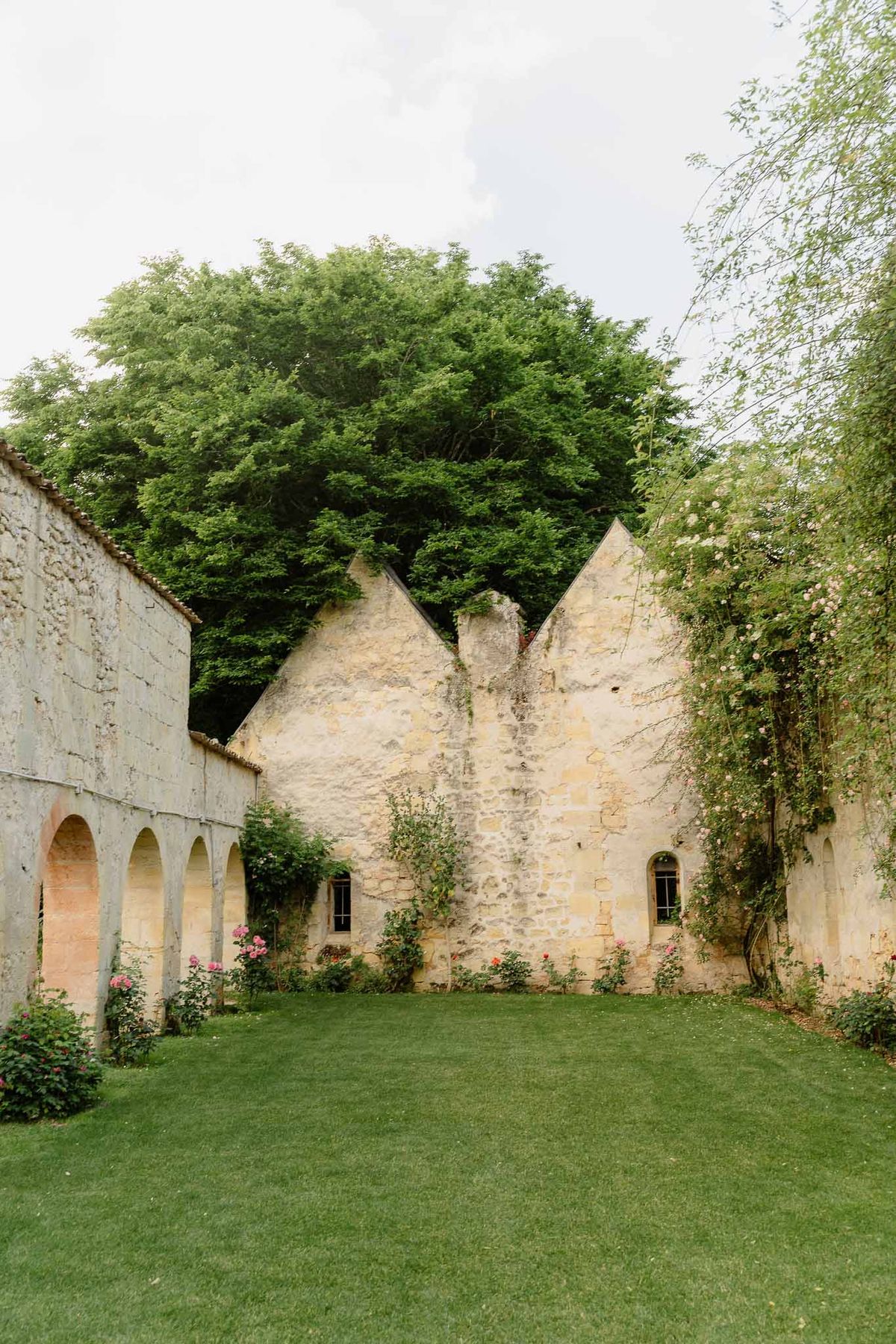Limestone walled courtyard with pointed gables, covered arcade, and pink climbing roses on lawn