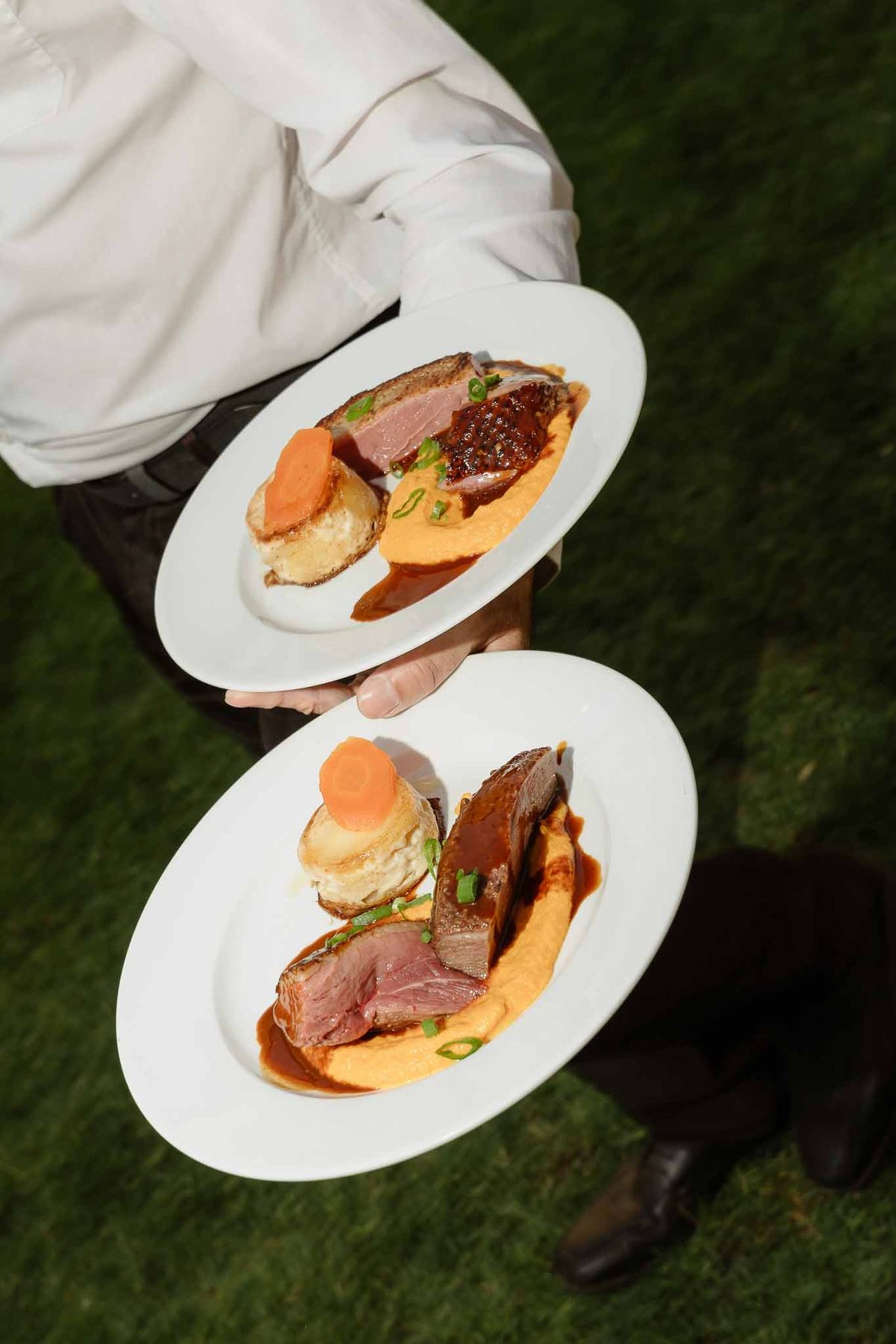 Server carrying two plated duck breast dishes with orange puree potato gratin and reduction sauce on white oval plates