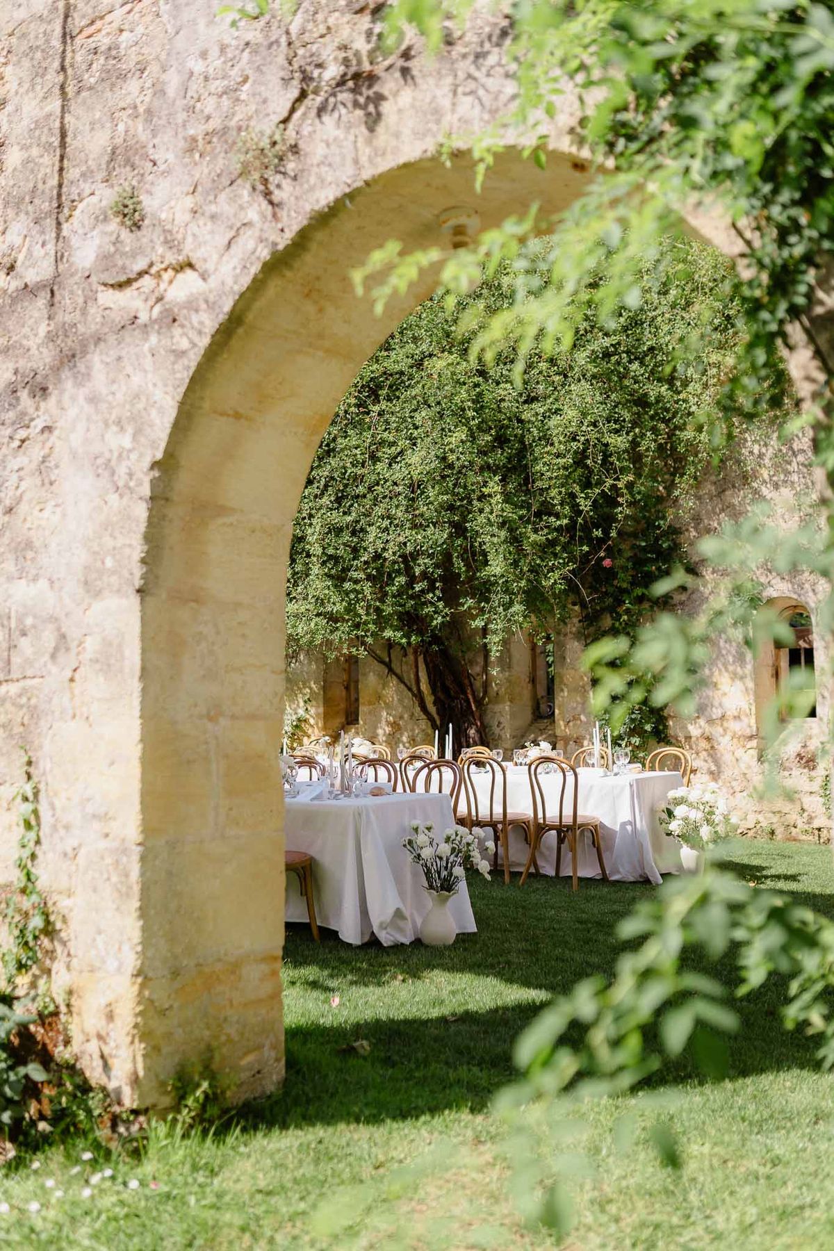 Outdoor reception tables with white linen and rattan chairs framed through stone archway with foliage