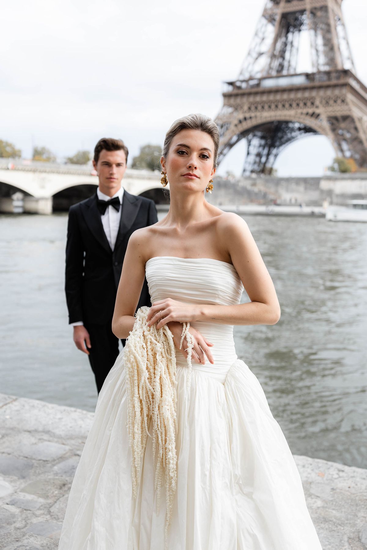 Bride holding a fringed bouquet beside the groom by the Seine in Paris