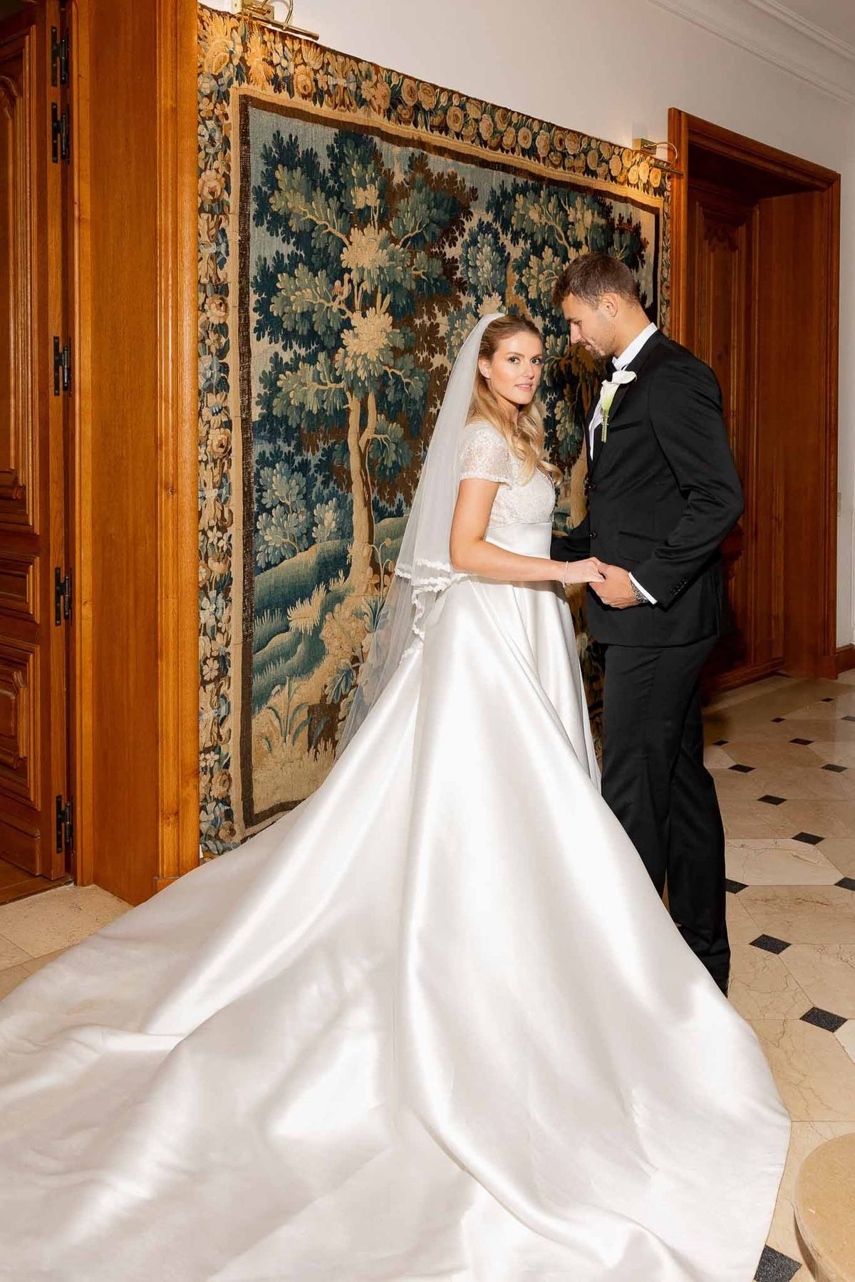 Bride and groom holding hands beside tapestry on marble floor at Château Comtesse Lafond