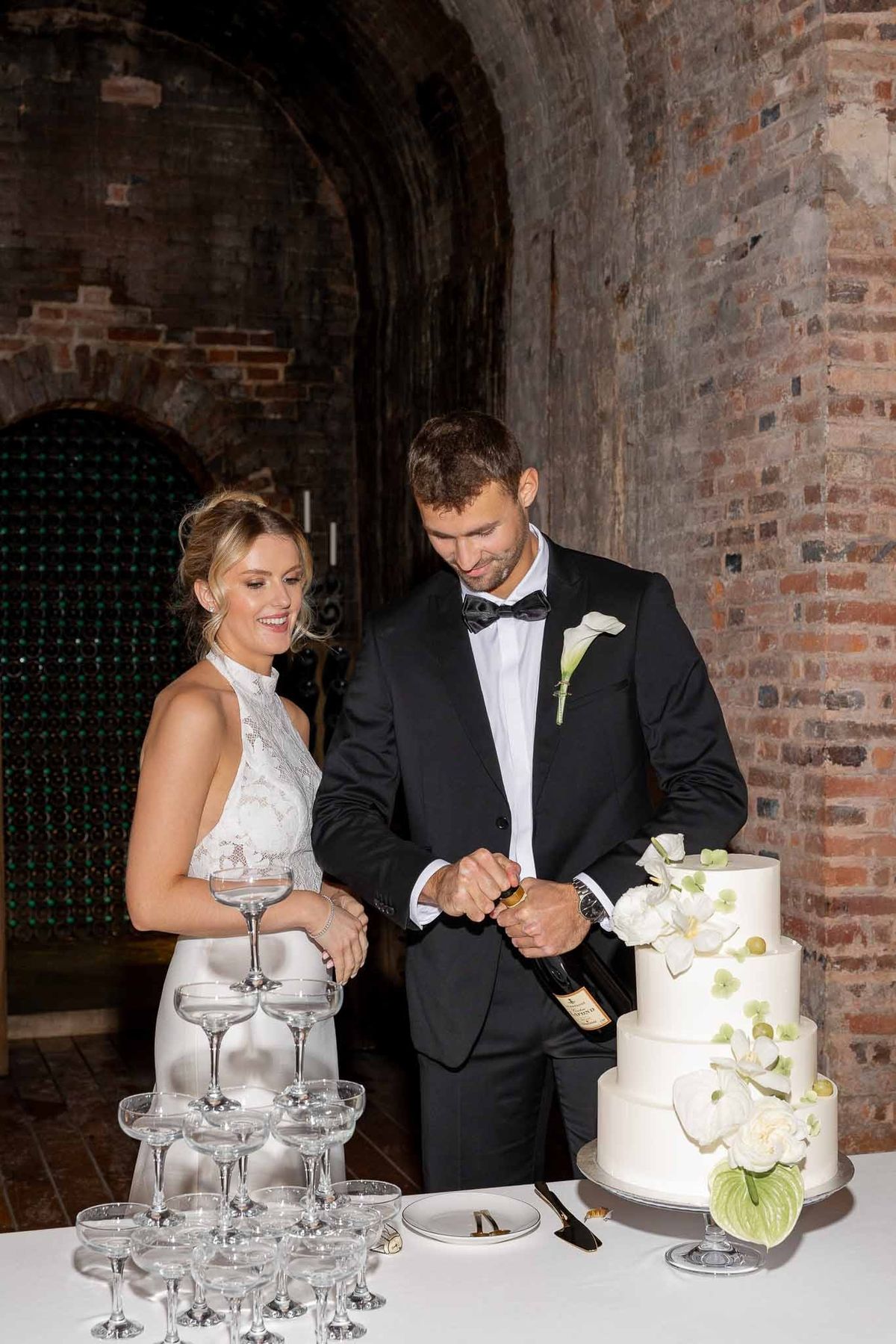 Couple opening champagne beside cake and glass tower in wine cellar at Château Comtesse Lafond