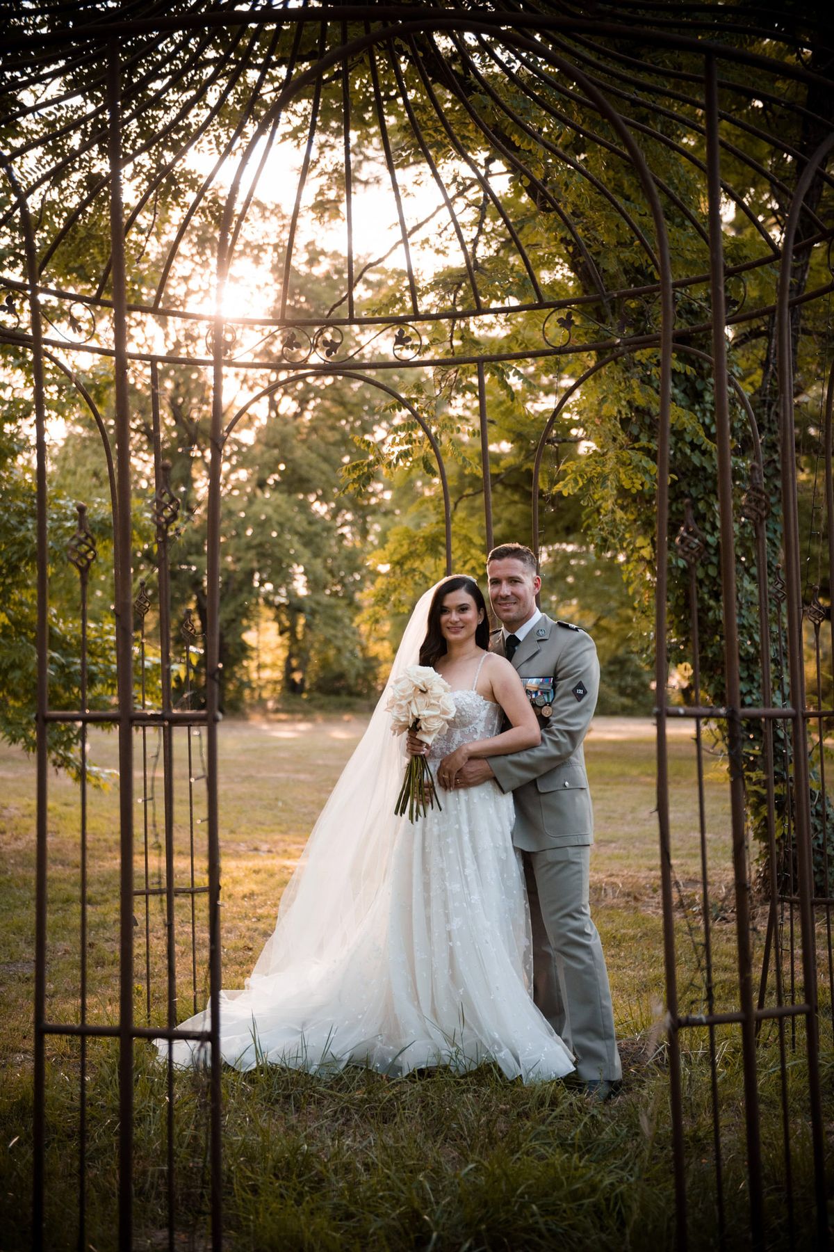 Couple in ivory ballgown and sage military uniform framed by wrought-iron gazebo with cream rose bouquet at golden hour