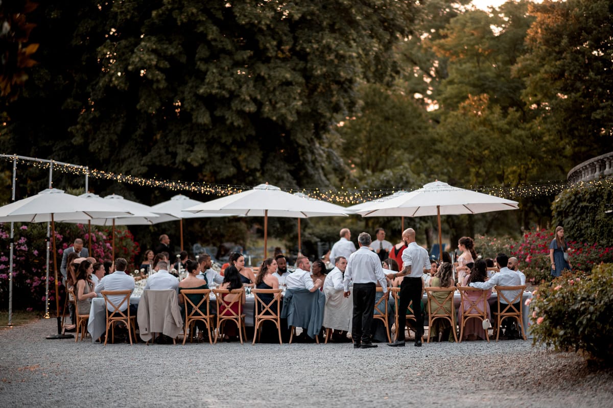 Thirty-plus guests at long tables under white parasols and fairy lights in formal garden at golden hour