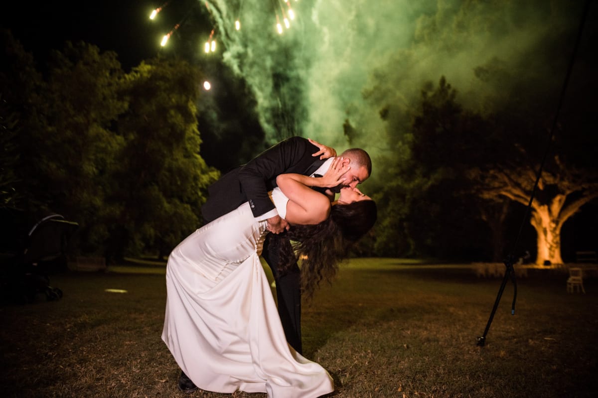 Bride and groom share a dip kiss outdoors at night with fireworks and golden sparks behind them