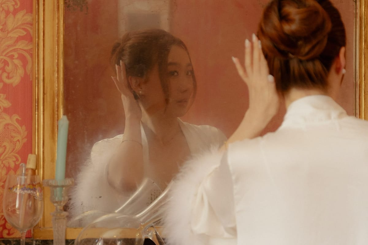 Bride in white satin robe adjusting earring before gold-framed antique mirror in rose-red damask wallpapered room