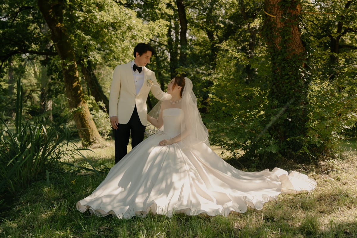 Bride seated in ballgown with veil as groom in cream dinner jacket rests hand on her head in wooded garden