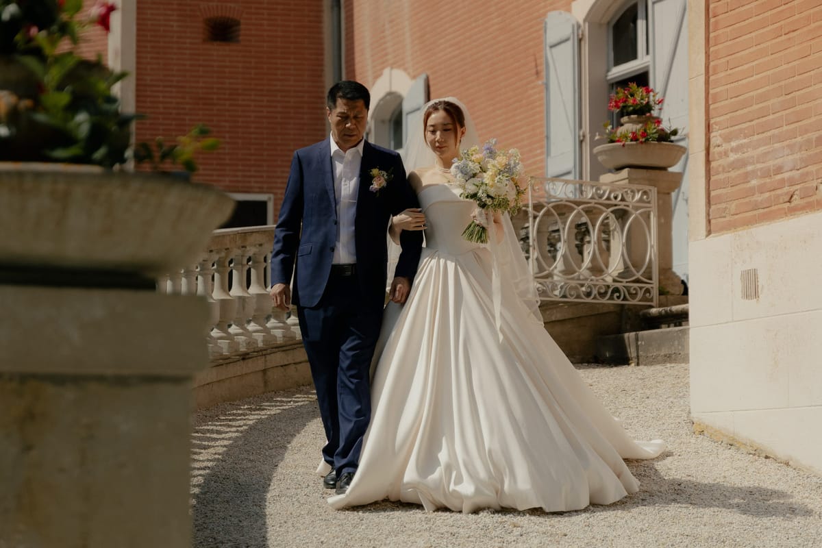 Bride in off-shoulder ivory satin ball gown escorted by father along gravel path outside red-brick chateau