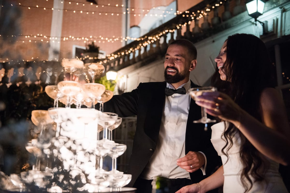 Couple pouring champagne into coupe tower with dry ice at nighttime courtyard reception