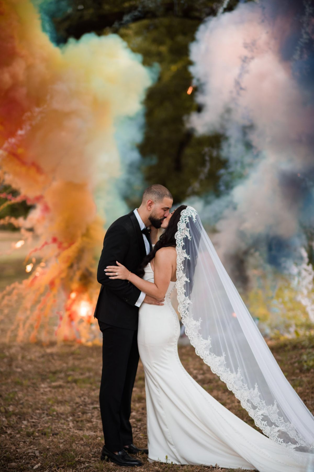 Bride in ivory mermaid gown and groom in tuxedo kissing amid orange, teal, and lavender smoke bombs