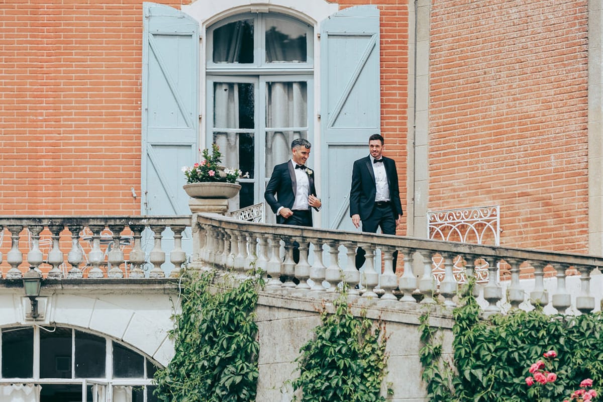 Groom and groomsman in tuxedos walk along stone balustrade terrace of red-brick chateau with pink roses