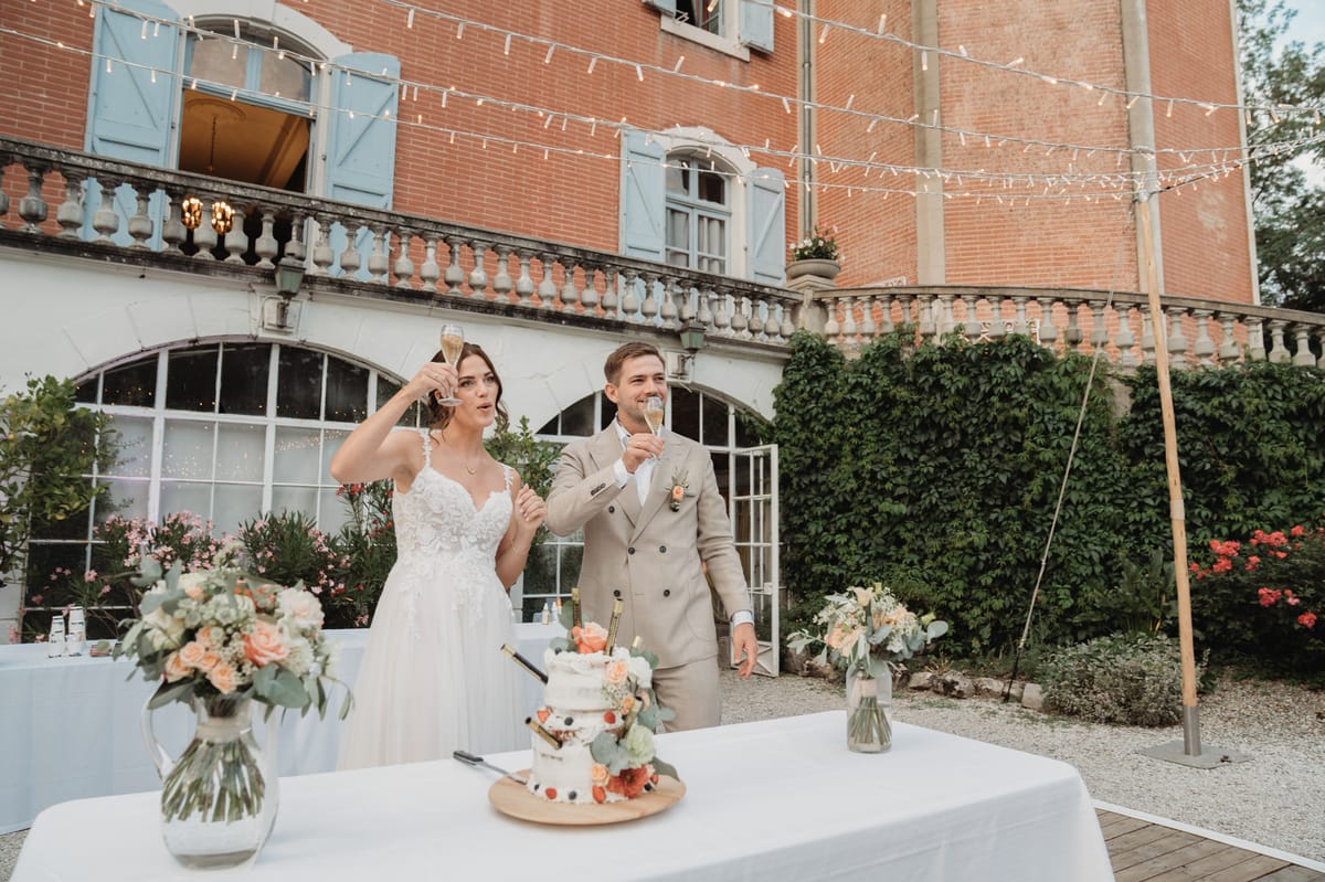 Couple toasting champagne by naked cake with peach roses at red-brick chateau with fairy lights at dusk