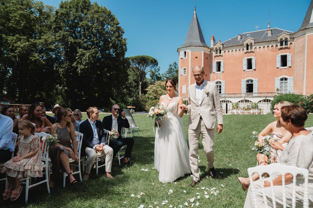 Bride walked down aisle by father at pink chateau with turret, white Chiavari chairs, and scattered petals