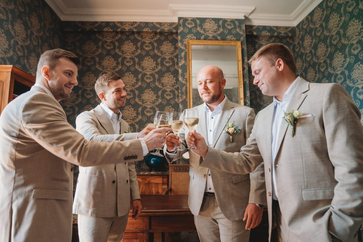 Four groomsmen in matching tan suits toast white wine in chateau room with teal damask wallpaper