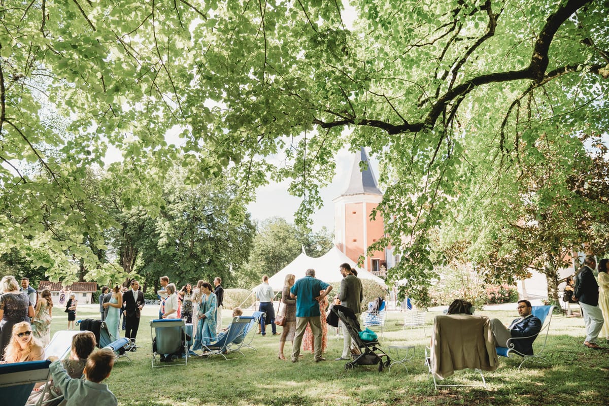 Thirty-plus guests mingling on lawn beneath mature trees with white marquee and brick chateau tower behind