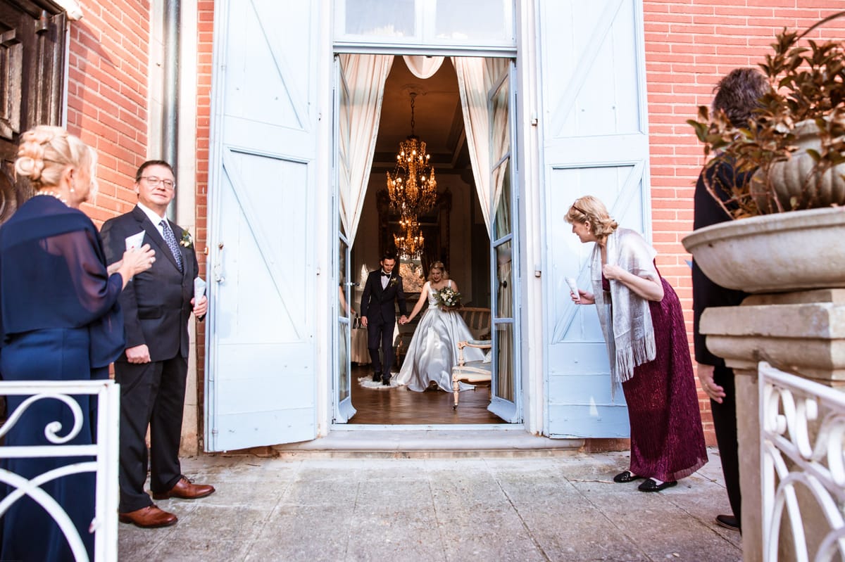 Couple exiting pale blue double doors of red brick manor as guests flank with confetti on stone terrace