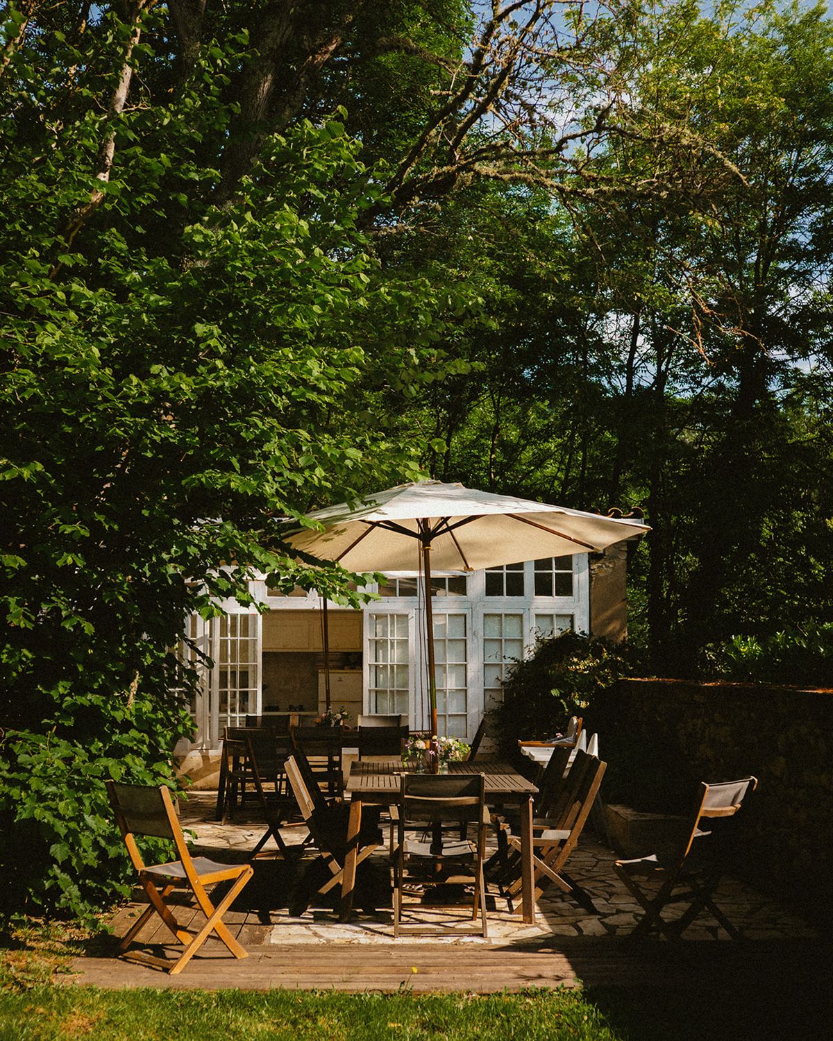 Stone-paved terrace with wooden tables, canvas chairs, and cream umbrella beside white orangerie